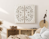 Two-panel carved wood wall art with symmetrical white floral and scroll patterns hanging on a light beige wall above a wooden cabinet and soft textiles.