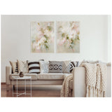 Two framed abstract floral canvases with soft pastel pink, green, and cream tones hung side by side above a beige sofa with black, white, and beige patterned pillows and a chunky knit throw.