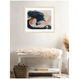 Abstract art with sweeping brushstrokes in deep blue, beige, and burnt orange colors framed in light wood, hanging above a white bench with a beige fur throw and a brown bag below.
