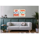 Two framed canvases with stylized wave graphics and bold text reading 'LA MER' and 'HIGH TIDE Surf Club' hang above a light gray tufted sofa with a round cushion in a modern living room.