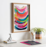 Abstract framed artwork with layered, curved brushstrokes in vibrant pink, blue, green, and orange hues, displayed on a white wall above a desk with a plant, cat figurine, and notebooks.