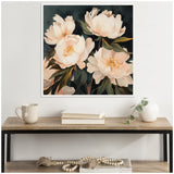 Square framed canvas painting of four soft pink peonies with green leaves on a dark background, hanging above a wooden table with a beige mug, books, and a small white vase.