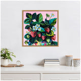 Square framed painting of ripe red and green strawberries with white blossoms and dark green leaves on a pink background, hanging on a white wall above a white shelf with a plant, books, and decorative containers.
