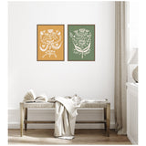 Two framed canvas prints with white floral and bird motifs, one on ochre background and the other on olive green, hung side by side above a wooden bench with light fabric draped