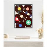 Black vinyl records with colorful center labels in yellow, pink, red, blue, green, and beige, arranged in a black frame on a white wall above a minimalist white table.