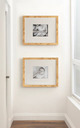 Two square picture frames with light wood grain and white mats hang vertically on a white wall, displaying black-and-white photos of a baby and a child.
