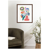 Framed whimsical floral art with stylized pink vase and colorful abstract flowers, hanging on a white wall above an olive green rounded armchair and a potted plant.