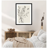 Black line drawing of protea flowers and leaves in a minimalist clear vase, framed in black, hanging above a wooden cabinet next to a cozy bed with beige and white linens.