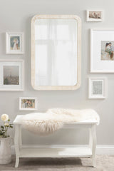 Rectangular wall mirror with rounded corners and a light wood frame hangs above a white wooden bench with a fluffy white throw, surrounded by framed photographs on a gray wall.