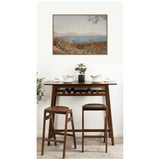 Warm wooden bar table with two matching stools, topped with white dishware and a small green plant, set against a white wall featuring a framed landscape painting.