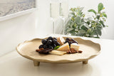 Round beige decorative tray with scalloped edges holding black grapes, assorted cheeses, crackers, almonds, and chocolate on a white surface near a green leafy plant and two champagne flutes.