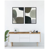 Two framed abstract canvases featuring overlapping rounded shapes in muted greens, grays, and black, hanging above a white and wood minimalist sideboard with decorative accents.