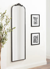 Tall rectangular wall mirror with arched top and ornate dark frame mounted next to two black-framed black-and-white photographs on white wall.