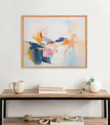 Abstract coastal print with soft pastel blocks and splashes in blue, peach, pink, and yellow framed in light wood, hanging above a wooden console with neutral decor.