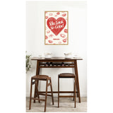 Framed wall art with a large red heart saying 'You Look So Good' surrounded by red lipstick kiss marks above a wooden bar table with two brown stools and white bowls.