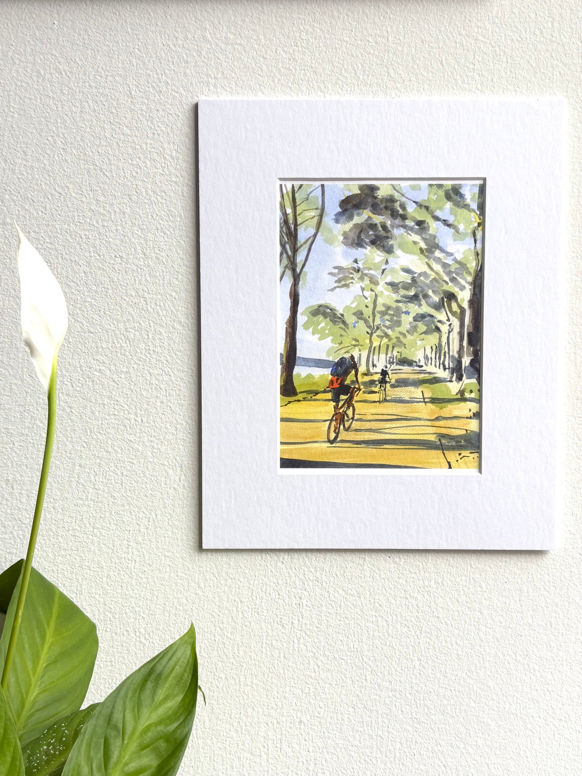 Watercolor painting of two bikers riding on a tree-lined path beside a river, with light filtering through the green leaves and casting shadows on the yellow path.