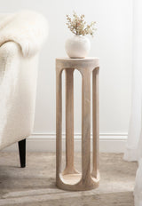 Tall cylindrical wooden side table with vertical cut-out arches and a smooth top holding a small white vase with dried flowers, placed beside a cream upholstered chair.