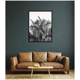 Black-and-white framed print of palm tree fronds hanging above a brown leather sofa against a dark green wall with a small wooden coffee table and decorative glass vase below.