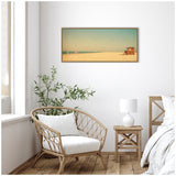 Framed rectangular wall art above a wicker armchair and wooden nightstand, depicting a sandy beach with a small lifeguard hut under a pale blue sky.