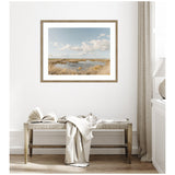 Framed photograph of a marshland under a partly cloudy sky with golden grasses and a small water body, hanging above a wooden bench with an open book and draped fabric.