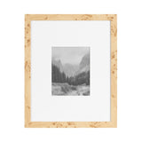 Light wood picture frame with natural burl patterns, showing a matte white border around a grayscale landscape photo of pine trees and mountains in mist.