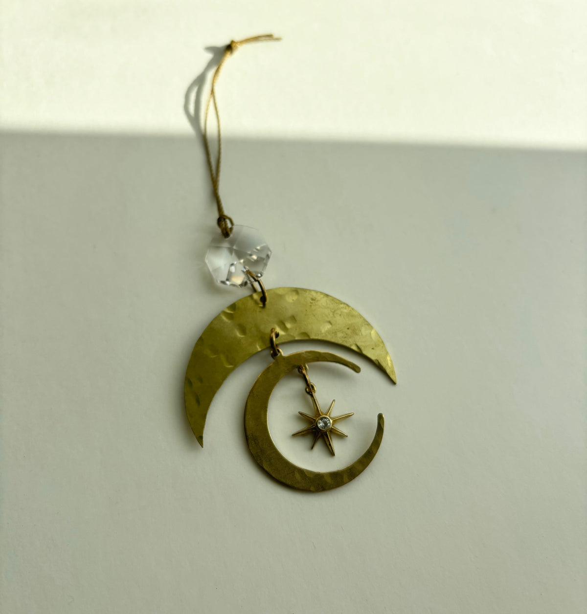 Brass crescent moon ornament with hammered texture, featuring a small star charm with a vintage glass crystal at the top, hanging by a thin golden string.