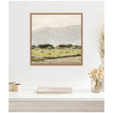 Framed canvas art of a muted mountain landscape with green fields and clusters of trees under a cloudy sky, displayed above a minimalist white shelf with dried flowers.