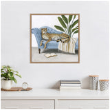 Framed canvas print of a cheetah lounging on a tufted blue chaise with a white fringed throw and a tall leafy plant behind it, above a stack of books on a white desk.