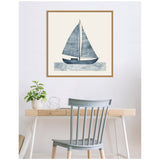 Framed canvas print of a navy blue sailboat with striped sails above stylized waves, hanging above a light wood desk with a plant and decorative wire orb, and a pale blue chair.