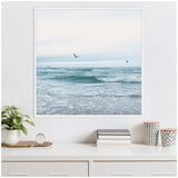 Square framed canvas of a calm ocean scene with gentle waves and three birds flying above under a soft, pale sky.
