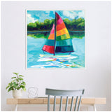 Framed canvas painting of a sailboat with vibrant rainbow sails on blue water, green shoreline and cloudy sky in the background, hanging above a desk with a gray chair and plant.