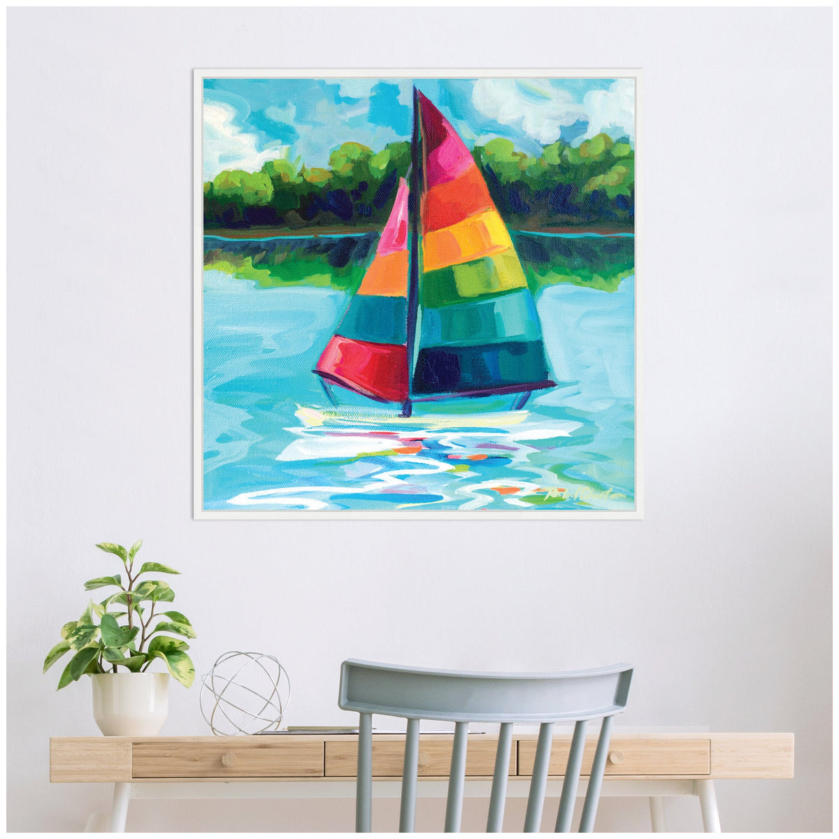Framed canvas painting of a sailboat with vibrant rainbow sails on blue water, green shoreline and cloudy sky in the background, hanging above a desk with a gray chair and plant.