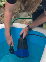 Person installing a black and blue submersible pump into a white tub filled with clear water outdoors, with stone wall and green garden hose in background.