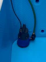 Blue and black cylindrical drain pump installed inside a textured blue container with a black power cable and a green hose attached.