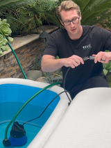 Man wearing glasses and a black RevX shirt attaches a hose clamp to a tube beside a blue inflatable cold plunge tub outdoors with plants and stone wall background.