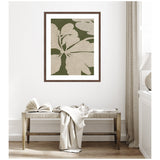 Framed botanical art print with large beige leaves on olive green background hanging above a light wood bench with an open book and beige fabric draped over it.