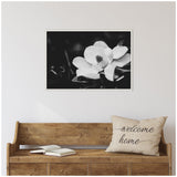 Black and white close-up of a magnolia flower with soft petals and a prominent center, framed and hung above a wooden bench with a beige 'welcome home' pillow.