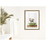 Framed print of a baby elephant sitting in a green clawfoot bathtub against a white paneled wall, displayed on a textured cream wall beside a towel rack and plant.