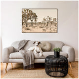Modern living room with beige sofa featuring a gray throw and round cushion, a black woven coffee table with a small green plant, and a framed Joshua Tree desert landscape hanging above.