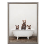 Three brown bear cubs sitting in a white vintage clawfoot bathtub on a wooden floor, with a light neutral background and a gray wooden frame.