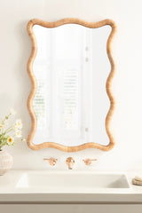 Rectangular mirror with a scalloped frame made of woven rattan, hanging above a white sink with brass faucet and handles in a light-colored bathroom.