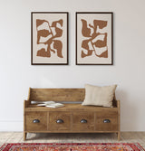 Two framed canvases side by side featuring abstract distorted shapes in brown and tan tones, hanging above a rustic wooden bench with a beige pillow and open book.