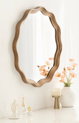 Wall-mounted round scalloped mirror with a smooth wooden frame in warm brown, reflecting sheer curtains and soft peach flowers in a white vase on a nearby surface.