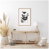 Black and white linocut artwork of three stylized birds, a flower, and foliage, framed in light wood on a white wall above a rustic wooden table with natural decor.