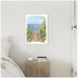 Framed watercolor painting of a wooden stairway leading through green foliage to a blue ocean under a pale sky, hanging on a white wall above a wooden bedside table.