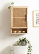 Light wood wall cabinet with woven rattan front panel and open cubby holding three small candles, mounted above a white toilet with trailing green plant and daisy artwork nearby.