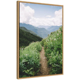 Narrow dirt trail flanked by lush green plants and white wildflowers, winding through a mountainous landscape under a partly cloudy blue sky.