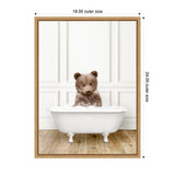 Cute brown bear cub sitting in a white vintage clawfoot bathtub filled with bubbles on a wooden floor, against a white paneled wall.