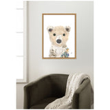 Framed canvas print of a stylized polar bear cub with large eyes, neutral fur tones, and a patch of birch tree bark and flowers near its shoulder, hanging above a dark gray armchair with a knitted cream throw.