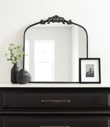 Black-framed arch mirror with ornate floral detail at the top, placed on a dark wood dresser with a gold handle, flanked by two ribbed black vases and a black-framed photo.